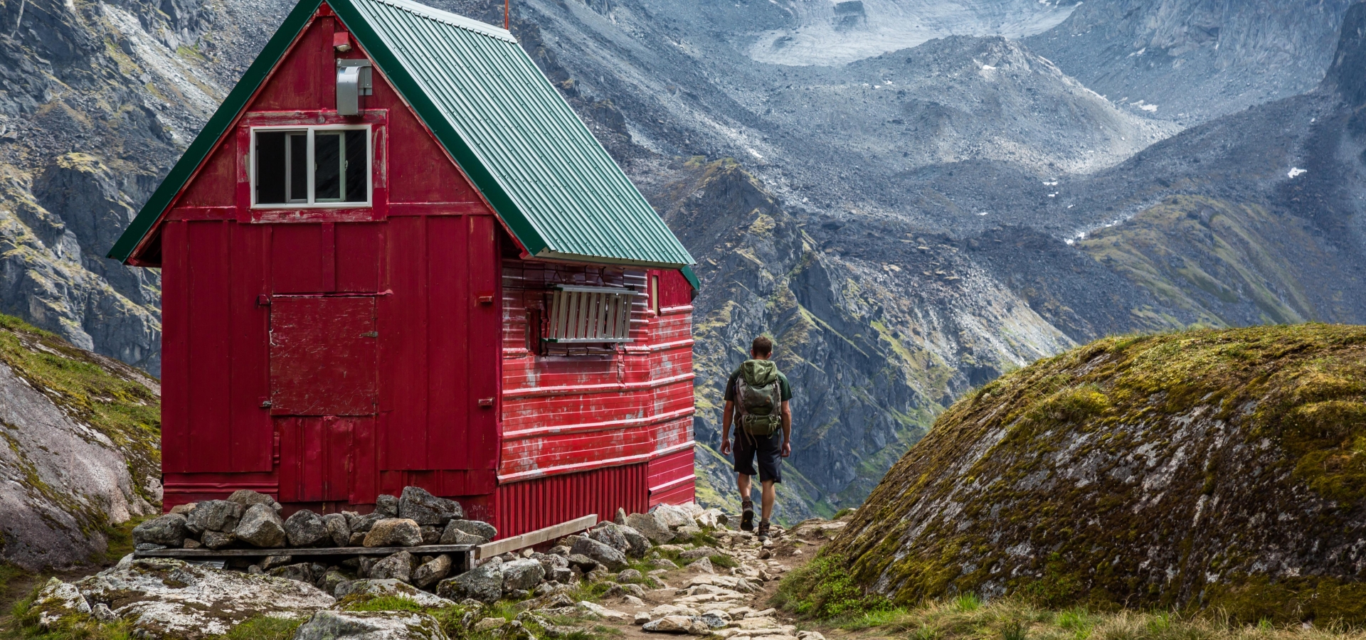 A man with a backpack out hiking walks up the trail to a red and green wilderness hut deep in the Talkeetna Mountains near Hatcher Pass, Alaska